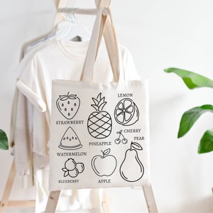 May include: A white canvas tote bag with black outlines of different fruits and their names. The fruits include a strawberry, pineapple, lemon, cherry, pear, watermelon, blueberry, and apple.