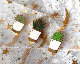 May include: Three enamel pins featuring potted succulents in different shades of green. The pins are on a white background with gold glitter and stars.