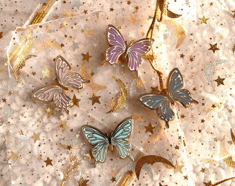 May include: Four enamel butterfly pins on a gold and white sparkly background. The butterflies are in shades of pink, blue, and purple.