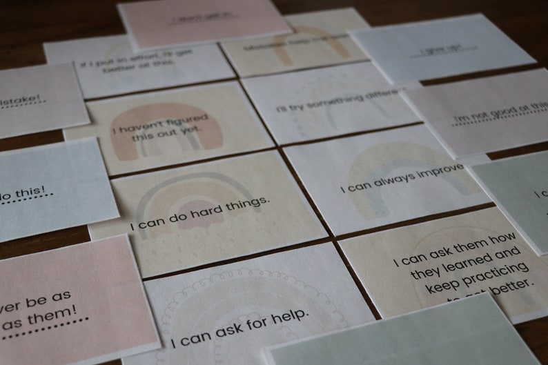May include: A collection of square cards with inspirational quotes and rainbow graphics. The cards are in various pastel colors, including pink, beige, and light green. Text on the cards includes phrases like "I can do hard things" and "I can always improve."