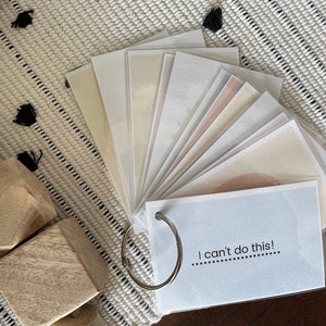 May include: A collection of color swatches fanned out on a white and black patterned surface. The swatches are in neutral tones and held together by a silver ring. The bottom swatch reads, "I can't do this!"