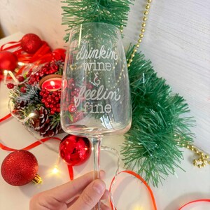 Può includere: Un bicchiere di vino in vetro trasparente con la scritta "drinkin wine & feelin fine" incisa sul lato. Il bicchiere è circondato da decorazioni natalizie rosse e verdi.