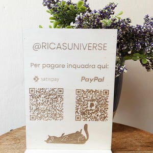 Può includere: Un cartello in legno bianco con il testo "@RICASUNIVERSE" e "Per pagare inquadra qui:" con due codici QR, uno per Satispay e uno per PayPal. Il cartello ha anche una piccola illustrazione di un gatto sdraiato sulla schiena.