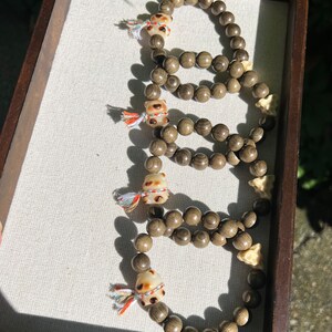 Könnte beinhalten: Fünf braune Holz-Armbänder mit kleinen, weißen und braunen Schweinchen-Anhängern. Jedes Armband hat einen bunten Quasten-Anhänger am Anhänger.