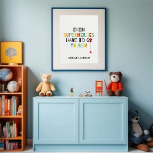 以下が含まれることがあります： カラフルな文字で「EVEN SUPERHEROES HAVE TO GO TO BED！」と書かれた額入りのプリント。プリントは、水色の壁に掛けられており、水色のキャビネットの上にあります。テディベア、おもちゃの車、その他のおもちゃがキャビネットにあります。