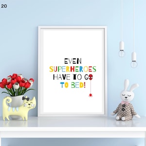 以下が含まれることがあります： カラフルな文字で「EVEN SUPERHEROES HAVE TO GO TO BED！」と書かれた額入りのプリント。プリントは、赤と白のチューリップが入った黄色の猫型の花瓶と、ぬいぐるみウサギが置かれた白いテーブルの上にあります。