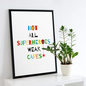 以下が含まれることがあります： カラフルな文字で「NOT ALL SUPERHEROES WEAR CAPES」と書かれた額入りのプリント。スーパーヒーローのロゴが特徴で、緑の葉を持つ鉢植えの植物の隣の白い棚に飾られています。