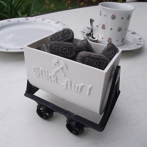 Könnte beinhalten: Ein weißer dekorativer Kohlenwagen gefüllt mit dunkelgrauen Briketts. Der Wagen hat schwarze Räder und einen schwarzen Rahmen. Auf dem Wagen steht der Schriftzug "Glück Auf!". Eine Blumetasse und ein Teller sind im Hintergrund.