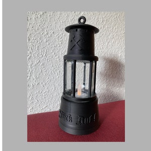 Op de afbeelding: Een zwarte metalen lantaarn met een glazen omhulling en een brandende kaars erin. De lantaarn heeft een houweel symbool bovenop en de woorden "Glück Auf!" onderaan.