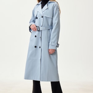 May include: A pale blue trench coat featuring a double-breasted design, a belt, and black buttons. The coat is worn with black trousers and heels. The model is wearing a black polo neck.