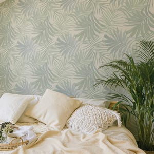 May include: A bedroom featuring wallpaper with a repeating sage green palm leaf pattern. The bed is covered with a cream-colored blanket and several pillows, including one with a macrame texture. A round woven tray holds two white mugs.