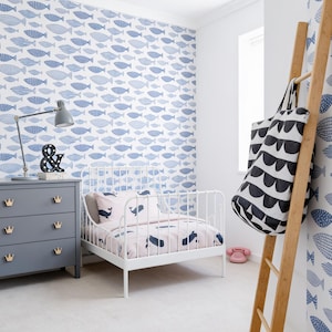 May include: A bedroom featuring white wallpaper with a blue fish pattern, a gray three-drawer dresser with gold crown-shaped knobs, a white metal bed frame with whale-patterned bedding, and a wooden ladder holding a black and white patterned bag.