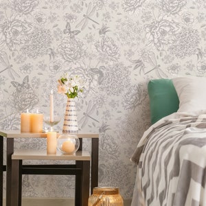 May include: A bedroom with a white and gray floral wallpaper, a bed with a white and gray striped blanket, a green throw pillow, and a set of nesting tables with candles and a vase of white flowers.