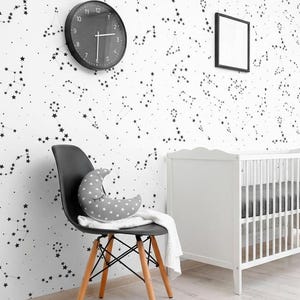 May include: A white wall covered in black constellation wallpaper patterns, featuring a black round wall clock, a black mid-century modern chair with a grey crescent moon pillow, and a white wooden crib with vertical slats and a grey mattress.