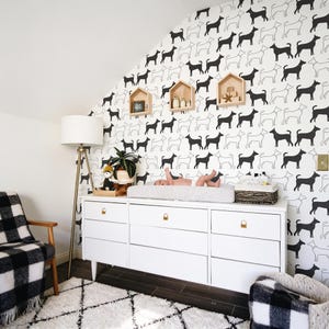 May include: A white dresser with six drawers and gold hardware, topped with a gray striped changing pad, a woven basket, and a small plant. The wall behind features a repeating black and white dog pattern wallpaper and three wooden house-shaped shelves.
