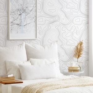 May include: A bedroom scene featuring white bedding with a cream-colored knit throw, a stack of books on a side table, and a wall covered in white wallpaper with a black topographic map line pattern and a framed map print hanging on the wall.