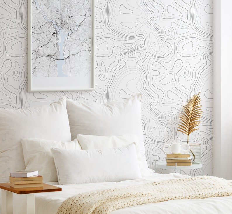May include: A bedroom scene featuring white bedding with a cream-colored knit throw, a stack of books on a side table, and a wall covered in white wallpaper with a black topographic map line pattern and a framed map print hanging on the wall.