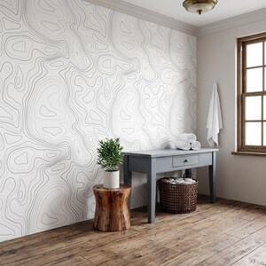 May include: A wall covered in white wallpaper featuring thin, black, wavy topographic contour lines. A small green potted plant sits on a cylindrical wooden stool next to a gray wooden console table with a woven basket and white towels.