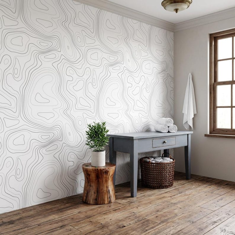May include: A wall covered in white wallpaper featuring thin, black, wavy topographic contour lines. A small green potted plant sits on a cylindrical wooden stool next to a gray wooden console table with a woven basket and white towels.
