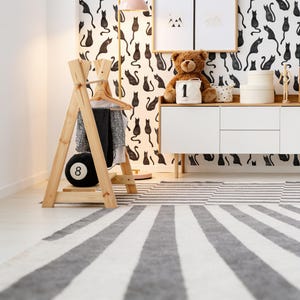 Puede incluir: Un perchero de madera clara en forma de A con una barra para colgar, un estante inferior y un coj&iacute;n negro con forma de bola de billar n&uacute;mero ocho. El perchero sostiene una percha de madera con una prenda superior estampada y pantalones negros.