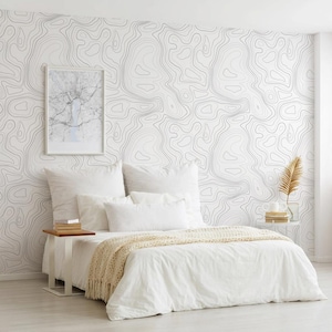May include: A bedroom featuring wallpaper with a black and white topographic map line pattern. The bed is dressed in white linens with a cream-colored knit throw blanket. A framed map print hangs on the wall above a small wooden side table.