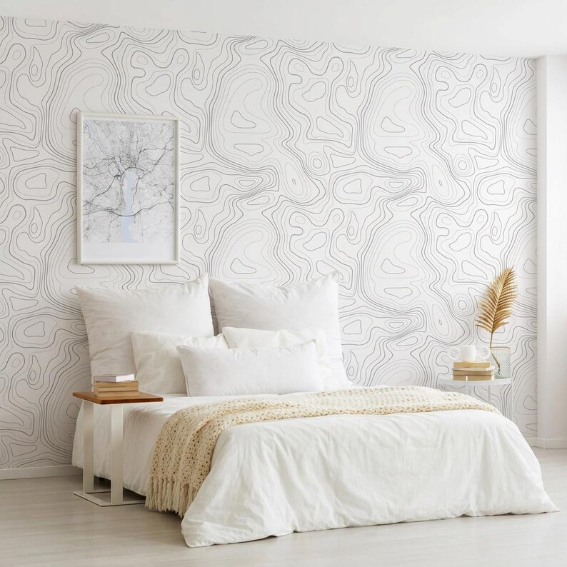 May include: A bedroom featuring wallpaper with a black and white topographic map line pattern. The bed is dressed in white linens with a cream-colored knit throw blanket. A framed map print hangs on the wall above a small wooden side table.