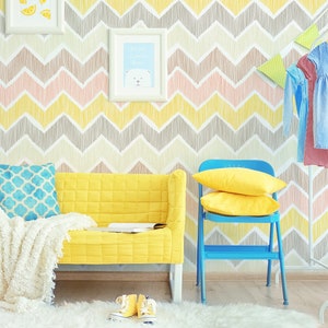 Puede incluir: Un sofá amarillo con una manta blanca y una almohada con estampado azul se encuentra frente a una pared con un patrón de chevron en tonos amarillos, rosas y grises. Una silla azul con almohadas amarillas se encuentra junto al sofá. Un perchero blanco con un vestido azul y rosa colgado está en el fondo.