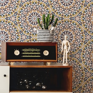 May include: A vintage wooden radio with a black face and silver knobs sits on a wooden shelf. The radio is in front of a white wall with a patterned wallpaper featuring blue, yellow, and brown floral designs. A small wooden figurine and a potted cactus are also on the shelf.