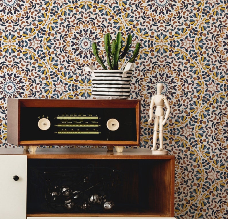 May include: A vintage wooden radio with a black face and silver knobs sits on a wooden shelf. The radio is in front of a white wall with a patterned wallpaper featuring blue, yellow, and brown floral designs. A small wooden figurine and a potted cactus are also on the shelf.