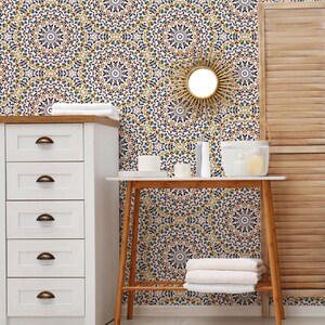 May include: A white dresser with four drawers and brass handles sits next to a wooden table with a white top. The table has a white ceramic vase with a white candle inside. The wall behind the table and dresser is covered in a patterned wallpaper with blue, yellow, and brown colors. A round mirror with a sunburst design hangs on the wall.