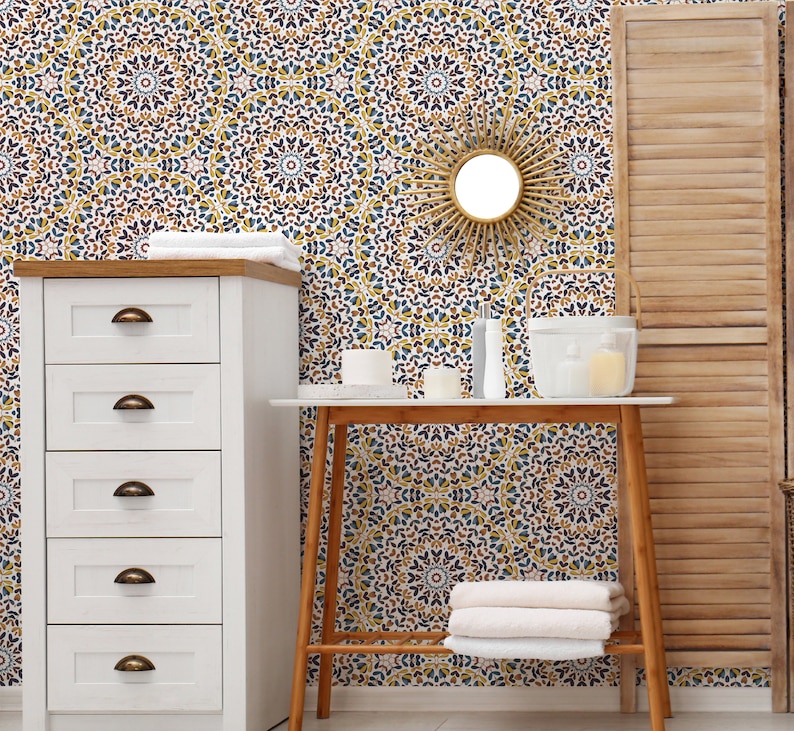 May include: A white dresser with four drawers and brass handles sits next to a wooden table with a white top. The table has a white ceramic vase with a white candle inside. The wall behind the table and dresser is covered in a patterned wallpaper with blue, yellow, and brown colors. A round mirror with a sunburst design hangs on the wall.