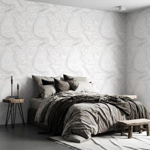 May include: A bedroom featuring wallpaper with a black topographic map line pattern on a white background. The bed is dressed in gray bedding with multiple pillows. A small wooden side table with two black candle holders stands to the left.