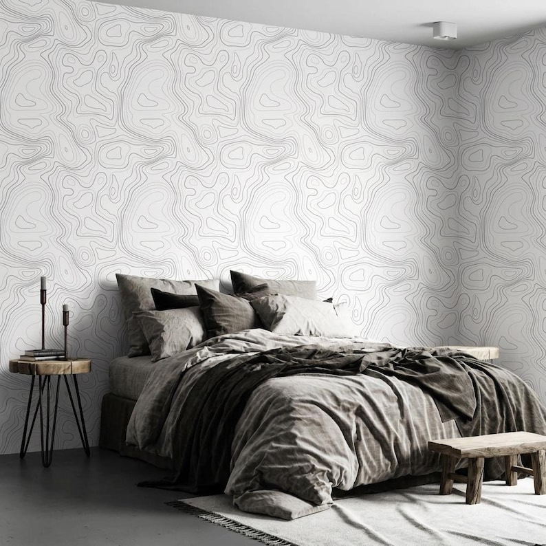 May include: A bedroom featuring wallpaper with a black topographic map line pattern on a white background. The bed is dressed in gray bedding with multiple pillows. A small wooden side table with two black candle holders stands to the left.