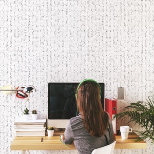 May include: A person wearing headphones sits at a desk in front of a computer. The desk is made of wood and has a lamp, a plant, and a cup on it. The wall behind the desk is covered in a white and gray patterned wallpaper.