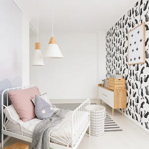 Puede incluir: Un marco de cama de metal blanco con una almohada rosa, un coj&iacute;n gris en forma de gota con una cara y una manta gris. La habitaci&oacute;n cuenta con dos l&aacute;mparas colgantes blancas con detalles en madera, una c&oacute;moda blanca y papel tapiz con patr&oacute;n de gatos negros.