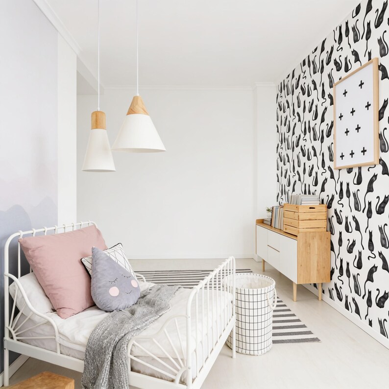 Puede incluir: Un marco de cama de metal blanco con una almohada rosa, un coj&iacute;n gris en forma de gota con una cara y una manta gris. La habitaci&oacute;n cuenta con dos l&aacute;mparas colgantes blancas con detalles en madera, una c&oacute;moda blanca y papel tapiz con patr&oacute;n de gatos negros.