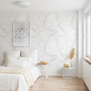 May include: A bedroom featuring wallpaper with a black and white topographic map line pattern. The bed is dressed in white linens with a textured throw blanket. A framed map print hangs on the wall above a small wooden side table and a wire basket.