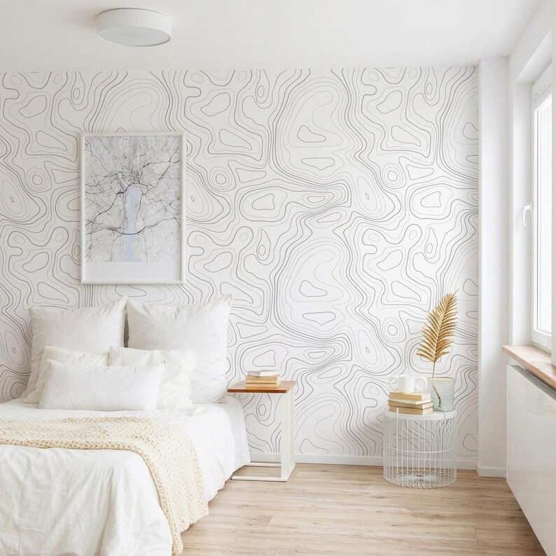 May include: A bedroom featuring wallpaper with a black and white topographic map line pattern. The bed is dressed in white linens with a textured throw blanket. A framed map print hangs on the wall above a small wooden side table and a wire basket.