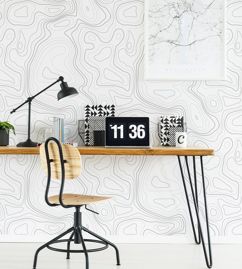 May include: A workspace featuring a wooden desk with black hairpin legs and a swivel chair with a light wood seat. The wall is covered in white wallpaper with black topographic contour lines. A laptop displays 11:36, next to a black desk lamp.