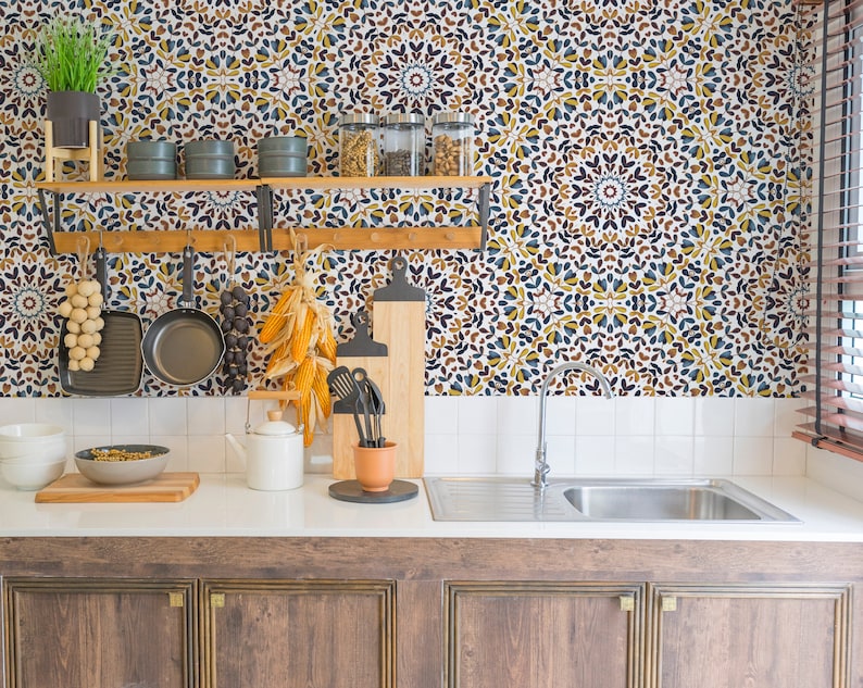 May include: A kitchen with a white tiled backsplash featuring a patterned wallpaper with blue, yellow, and brown colors. The wallpaper has a geometric design. The kitchen has a wooden countertop with a sink, a white teapot, and a bowl of food. There are wooden cabinets below the countertop.