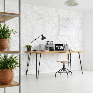 May include: A home office setup featuring a long wooden desk with black hairpin legs, a black task lamp, a laptop displaying the time 11:36, and a black swivel chair. The wall is covered in white wallpaper with thin black topographic contour lines.