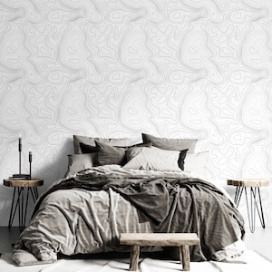 May include: A bedroom featuring wallpaper with a black and white topographic map line pattern. The bed is dressed in gray and charcoal bedding with multiple pillows. Two wooden side tables with black metal legs and a wooden bench are present.