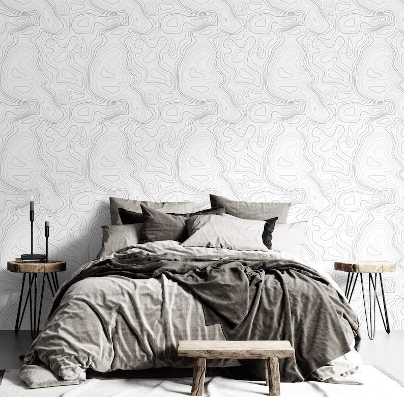 May include: A bedroom featuring wallpaper with a black and white topographic map line pattern. The bed is dressed in gray and charcoal bedding with multiple pillows. Two wooden side tables with black metal legs and a wooden bench are present.