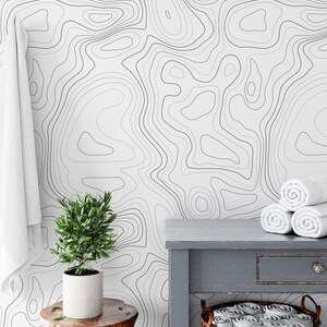 May include: A white wall covering featuring a repeating dark gray topographic map line pattern. The design consists of fluid, irregular contour lines that create a layered, map-like aesthetic across the entire surface of the wall.