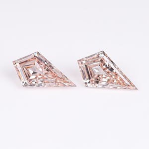 May include: Two loose kite-shaped gemstones with a pale peach hue. Each stone features a faceted surface with a central point and geometric cuts that reflect light, creating a symmetrical appearance with sharp edges and a polished finish.