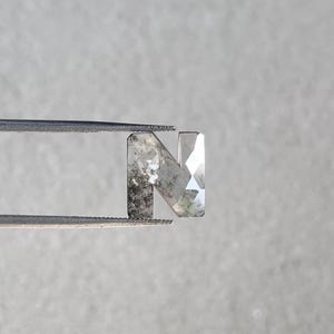 Pode incluir: Uma gema facetada, cinza claro, em forma da letra "N" é segurada por uma pinça. A gema tem uma mistura de áreas claras e nebulosas, com um corte geométrico no lado direito. A pinça é prateada e o fundo é cinza.