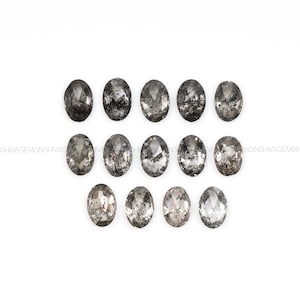 Op de afbeelding: Een verzameling van 14 ovale, zwarte diamanten met een ruwe, ongeslepen afwerking. De diamanten zijn in drie rijen op een witte achtergrond gerangschikt.