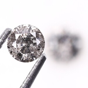 May include: A round, salt and pepper diamond held by metal tweezers against a white backdrop. The diamond has a speckled appearance with grey and black inclusions. Another diamond is blurred in the background.