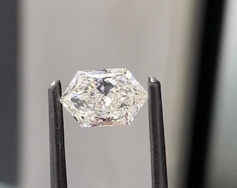 May include: A faceted, hexagonal-cut diamond held by black tweezers. The diamond has a brilliant, clear appearance, reflecting light. The gemstone is the focal point, showcasing its intricate facets and overall clarity. The background is a soft, neutral gray.