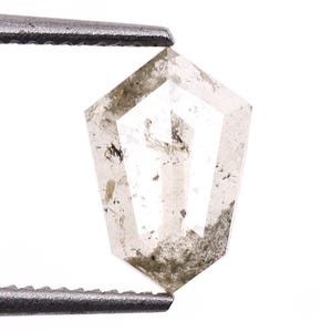 May include: A clear, shield-cut diamond with inclusions, held by a pair of metal tweezers. The gemstone has a multifaceted design with a light gray and black color scheme. The diamond's shape is a modified pentagon.